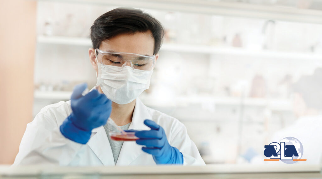 Scientist in lab coat, safety glasses, face mask, and gloves adding sample to petri dish