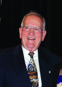 A headshot of a man with grey hair and glasses, wearing a suit and colorful tie