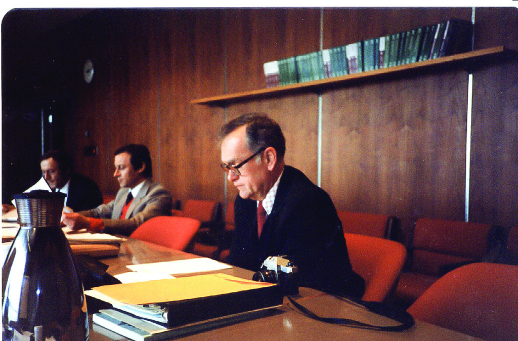 An old photo of a man with brown hair and glasses sitting in an orange chair at a long desk with business files in front of him.