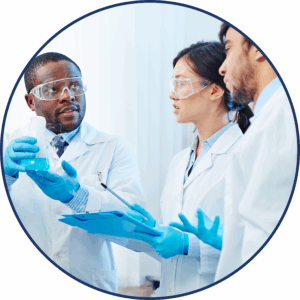 A woman and two men in white lab coats wear safety glasses and talk over a beaker with blue liquid and a clip board.