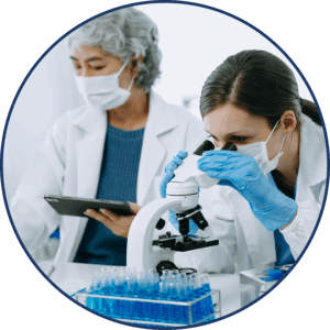 A scientist in a white lab coat, face mask, and rubber gloves looks into a microscope while another scientist in similar attire references a digital tablet